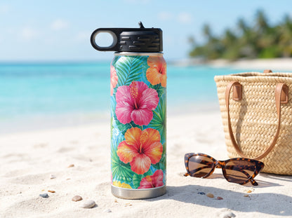 Tropical hibiscus water bottle standing on a sandy beach beside sunglasses and a woven tote.
